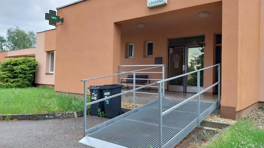 Vstup do lékárny v samostatné jednopodlažní budově s oranžovou fasádou a kovovým bezbariérovým nájezdem.